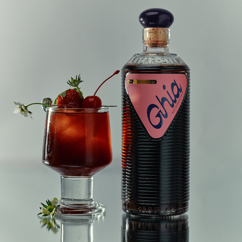 A bottle of Ghia non-alcoholic aperitif with a glass of cocktail, garnished with a cherry, strawberry, and flower. The bottle has a pink label with the word "Ghia.