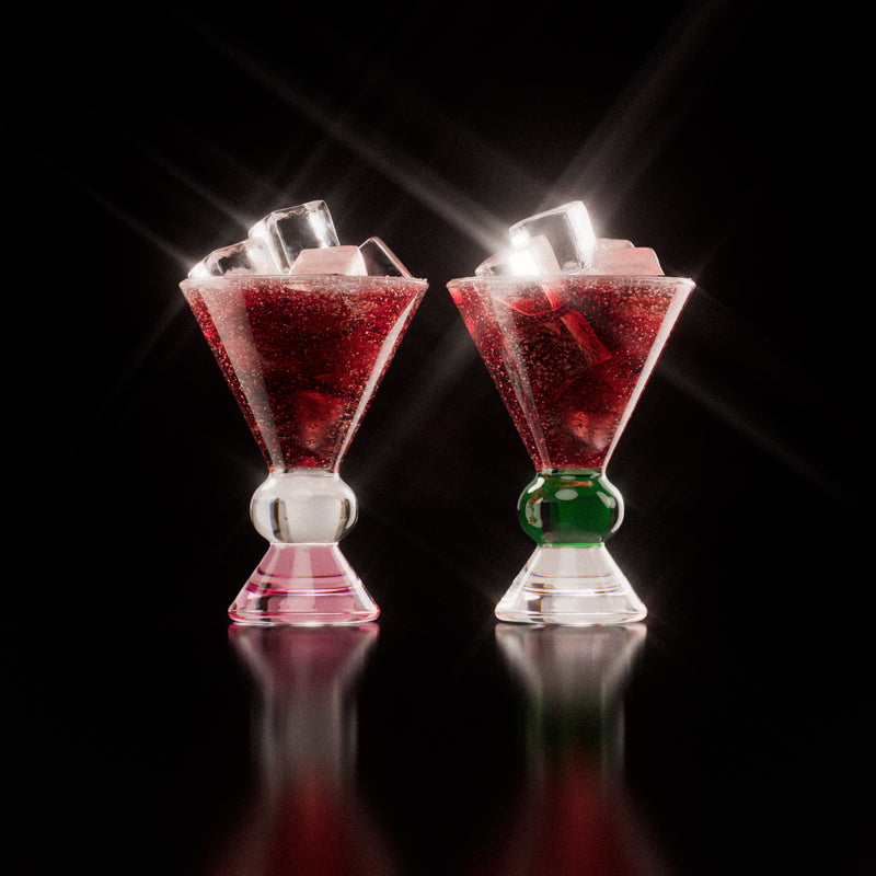 Two decorative glasses that have a pink, clear and green base, each containing a red drink with ice cubes, highlighting the distinctive totem-inspired shape of the glasses against a light background.