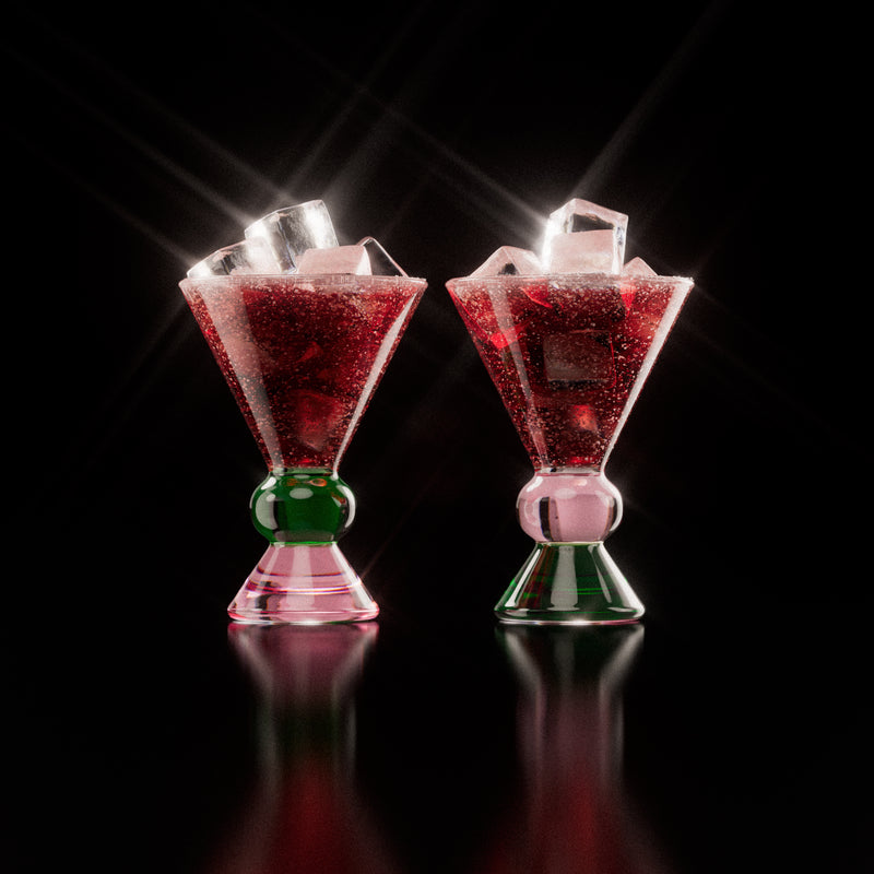 Two decorative glasses with a pink and green base, each containing a red drink with ice cubes, highlighting the distinctive totem-inspired shape of the glasses against a light background.