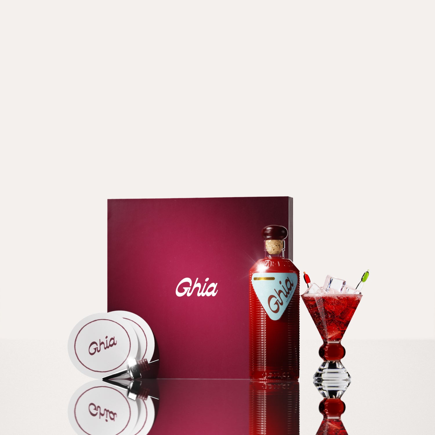 A Ghia non-alcoholic aperitif gift set with a burgundy box, paper coasters, 1 glass bottle of Original Apéritif, and a decorative cocktail glass that has a amber and clear base.