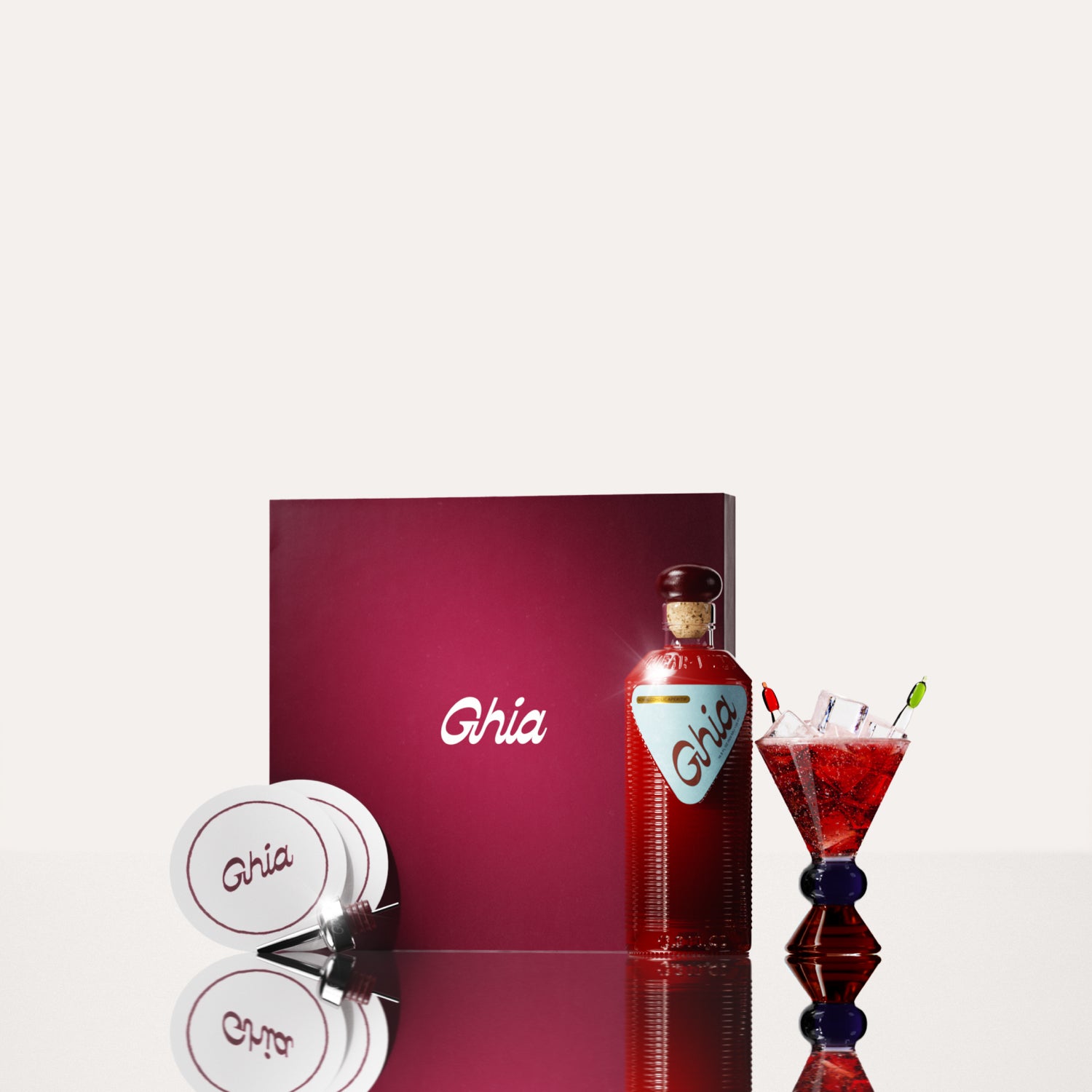 A Ghia non-alcoholic aperitif gift set with a burgundy box, paper coasters, 1 glass bottle of Original Apéritif, and a decorative cocktail glass that has a blue and amber base.