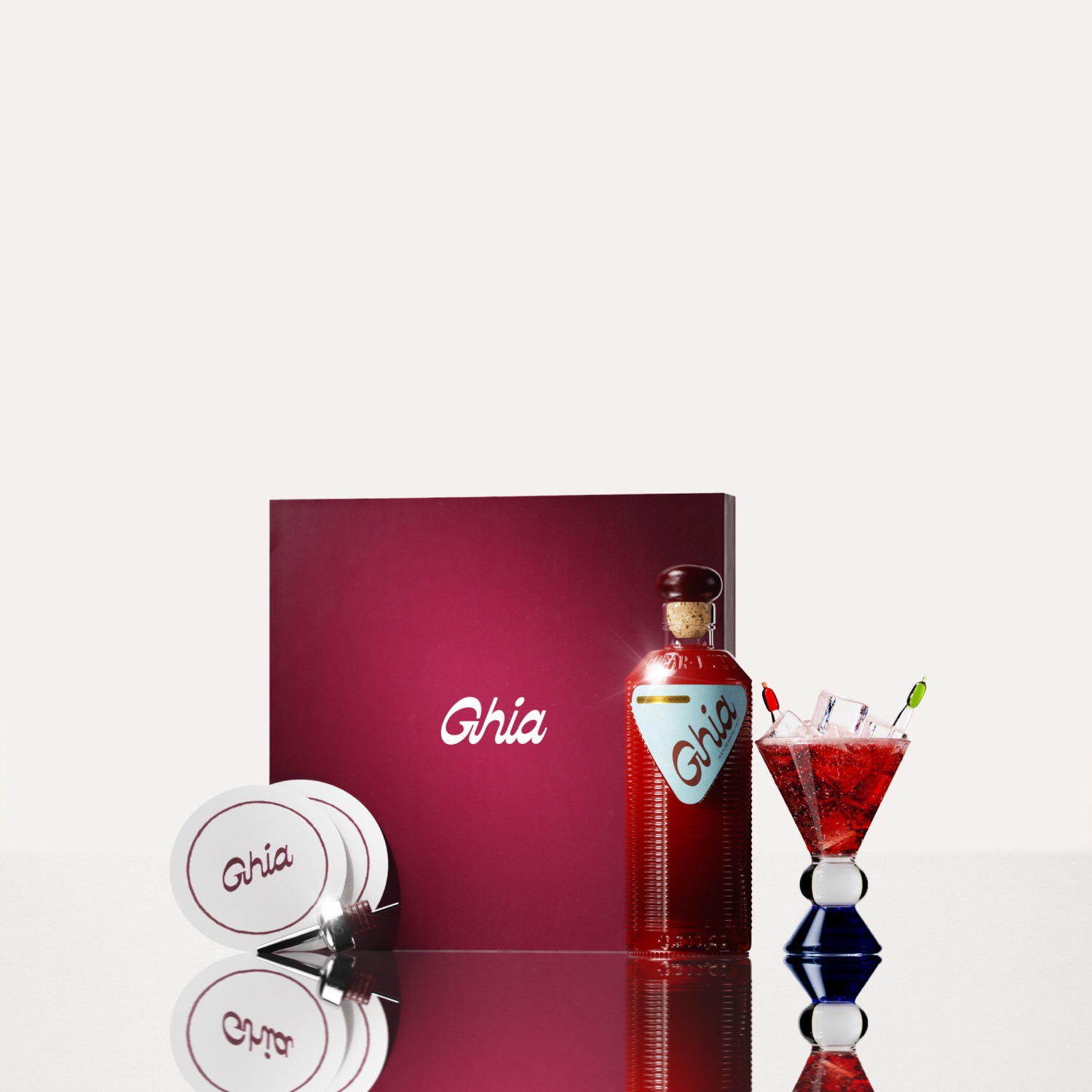 A Ghia non-alcoholic aperitif gift set with a burgundy box, paper coasters, 1 glass bottle of Original Apéritif, and a decorative cocktail glass that has a clear and blue base.