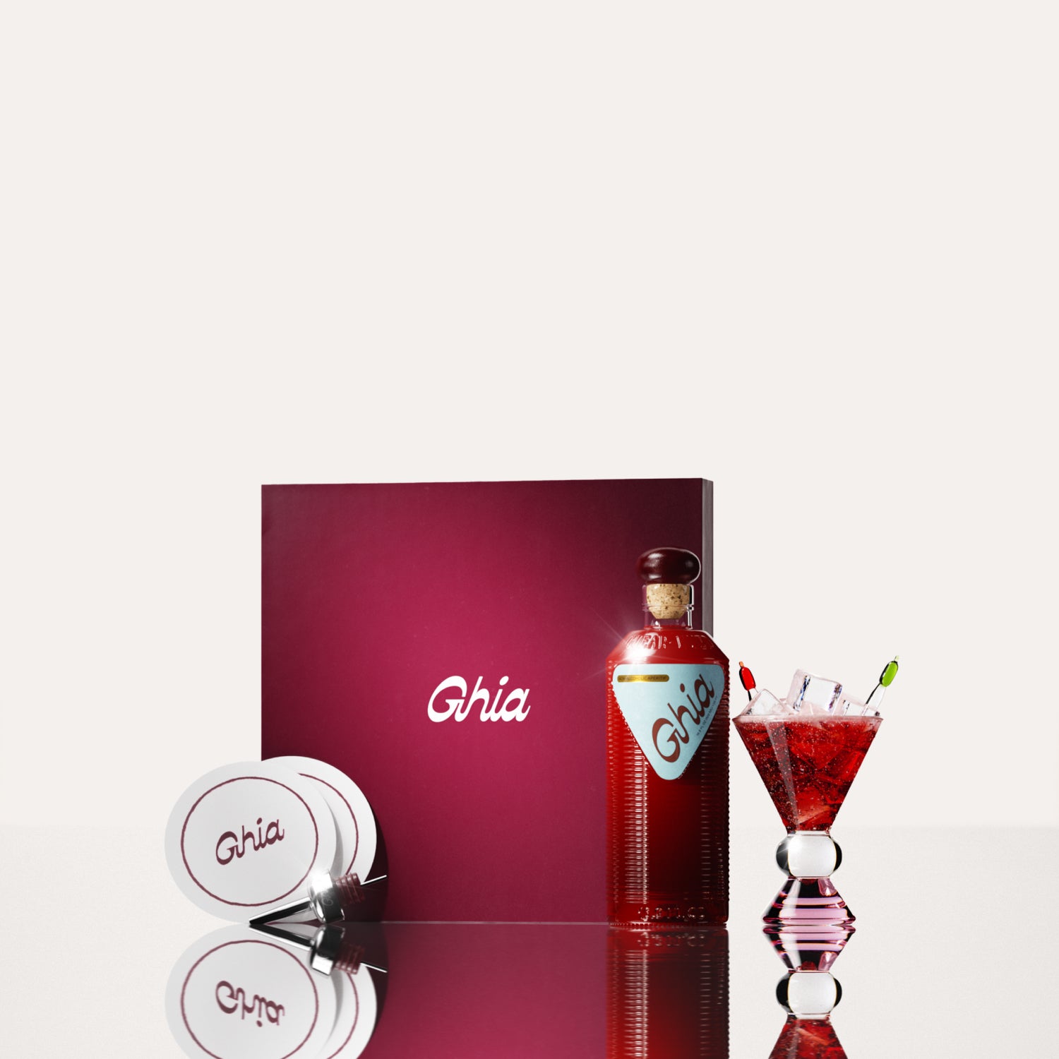 A Ghia non-alcoholic aperitif gift set with a burgundy box, paper coasters, 1 glass bottle of Original Apéritif, and a decorative cocktail glass that has a clear and pink base.