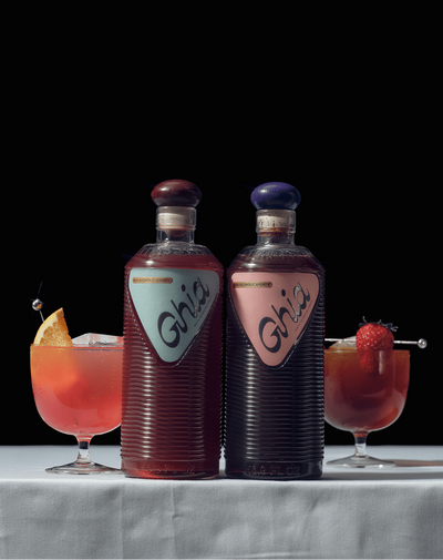 Two bottles of Ghia's with two cocktails on a marble surface against a black background