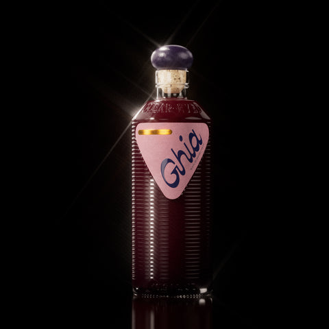 A Ghia non-alcoholic beverage in flavor Berry Apéritif clear bottle with ribbed glass, a purple cork top, and a pink label featuring bold branding on a reflective surface.