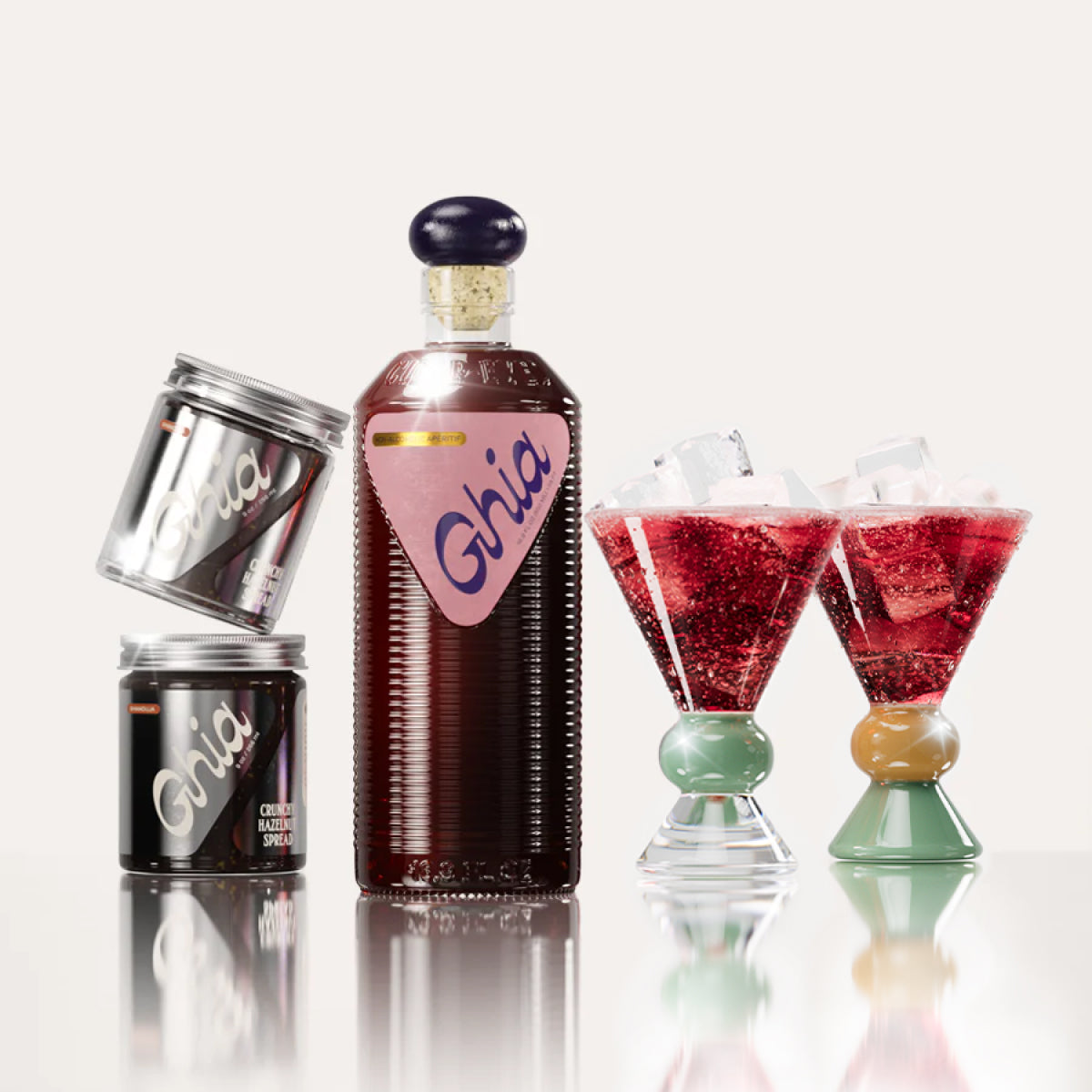 Deep-red non-alcoholic beverage in flavor Berry Apéritif in ribbed glass bottle with a pink label next to two jars of chocolate Hazelnut spread, and two cocktail glasses with a clear, jade and sand base.