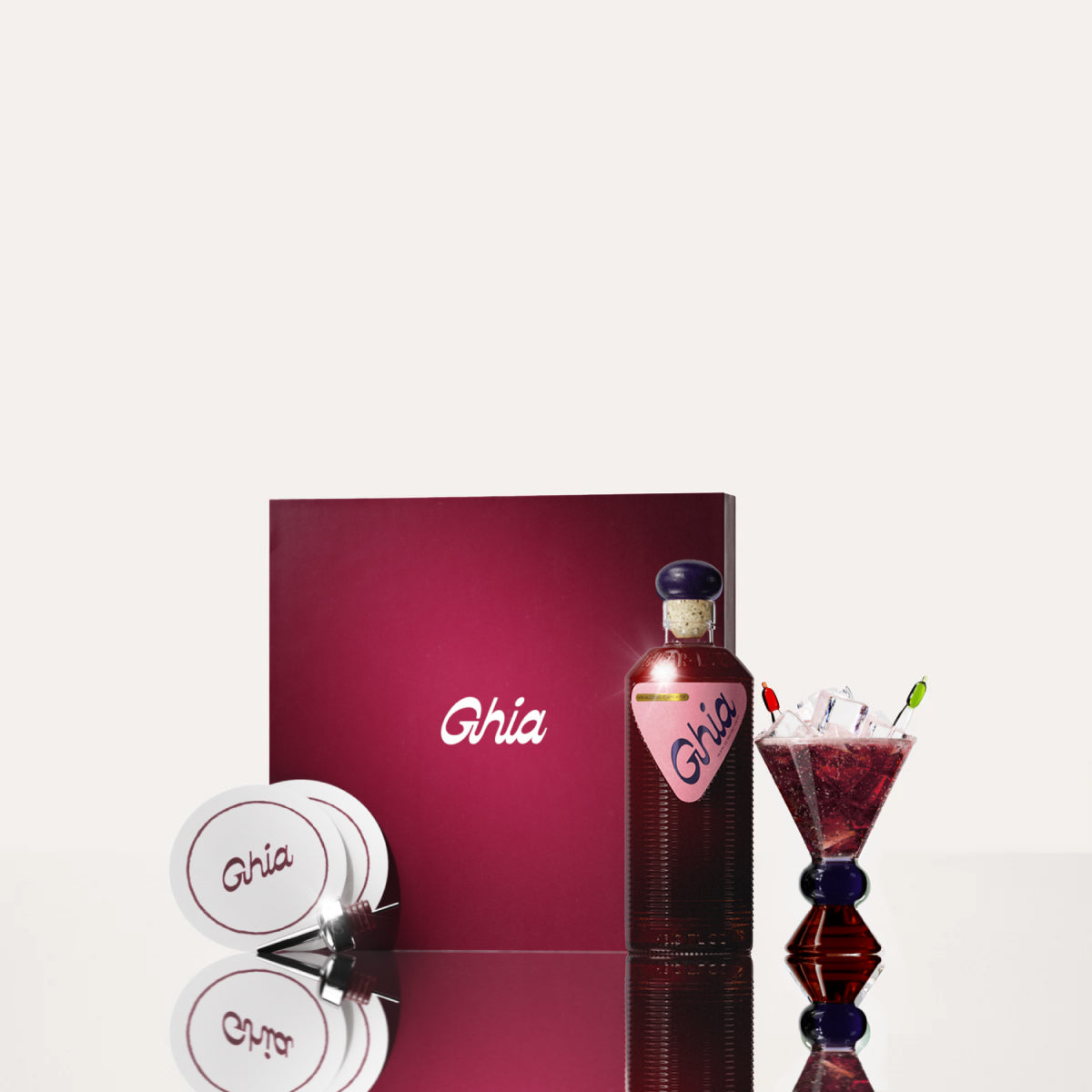 A Ghia non-alcoholic aperitif gift set with a burgundy box, paper coasters, 1 glass bottle of Berry Apéritif, and a decorative cocktail glass that has a blue and amber base.