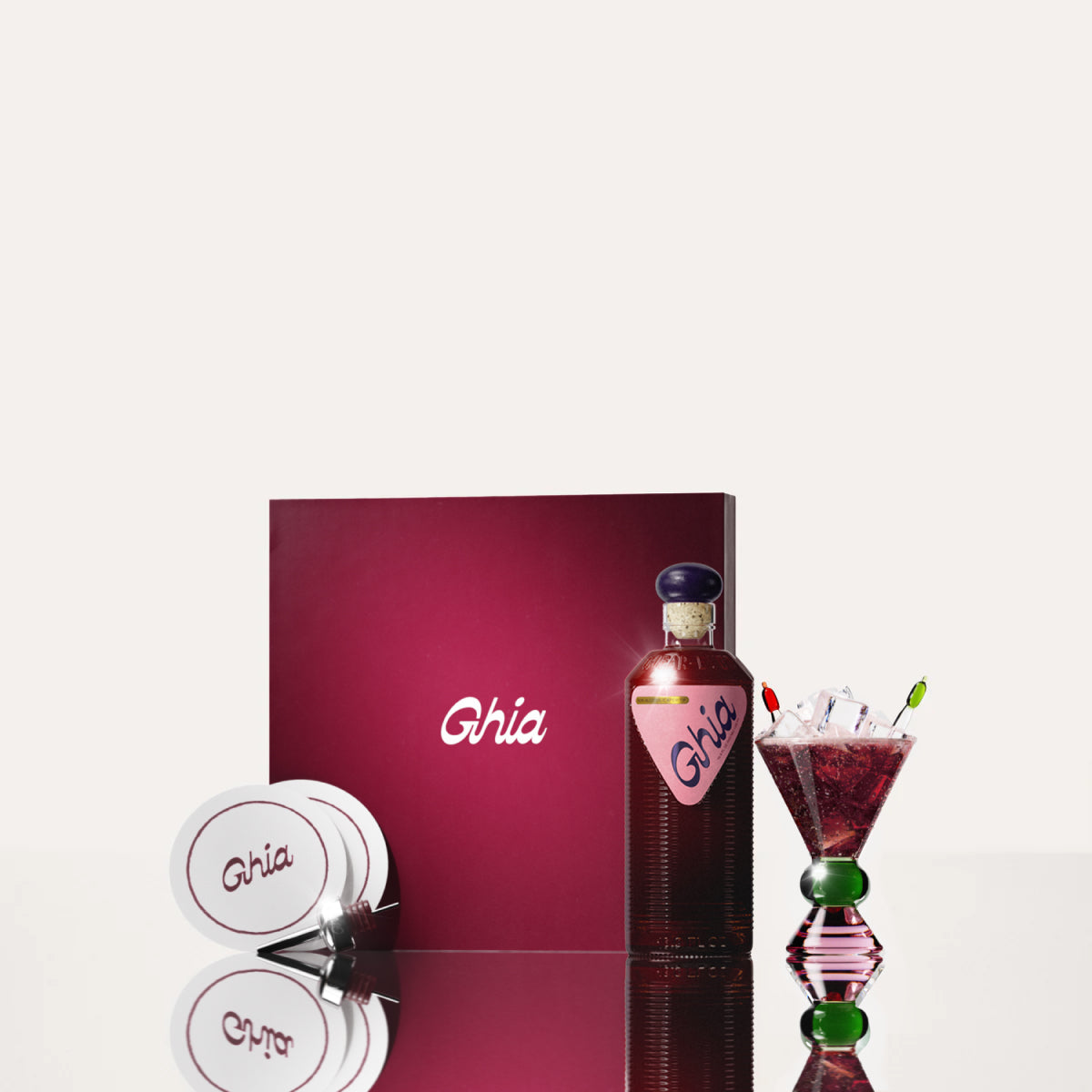 A Ghia non-alcoholic aperitif gift set with a burgundy box, paper coasters, 1 glass bottle of Berry Apéritif, and a decorative cocktail glass that has a green and pink base.