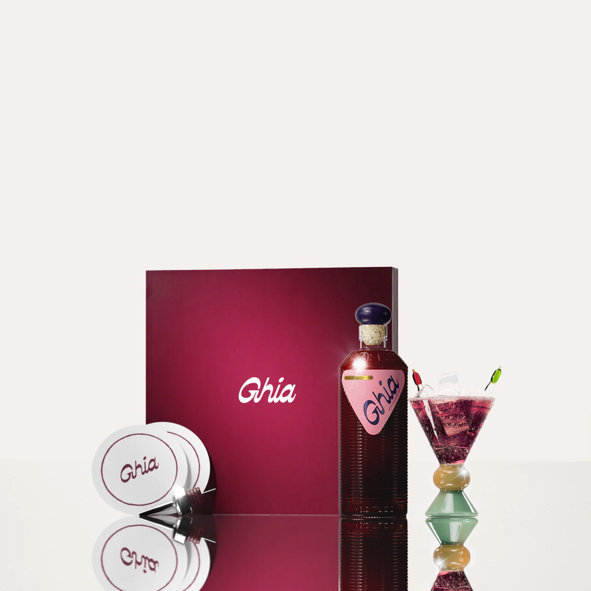 A Ghia non-alcoholic aperitif gift set with a burgundy box, paper coasters, 1 glass bottle of Berry Apéritif, and a decorative cocktail glass that has a sand and jade base.