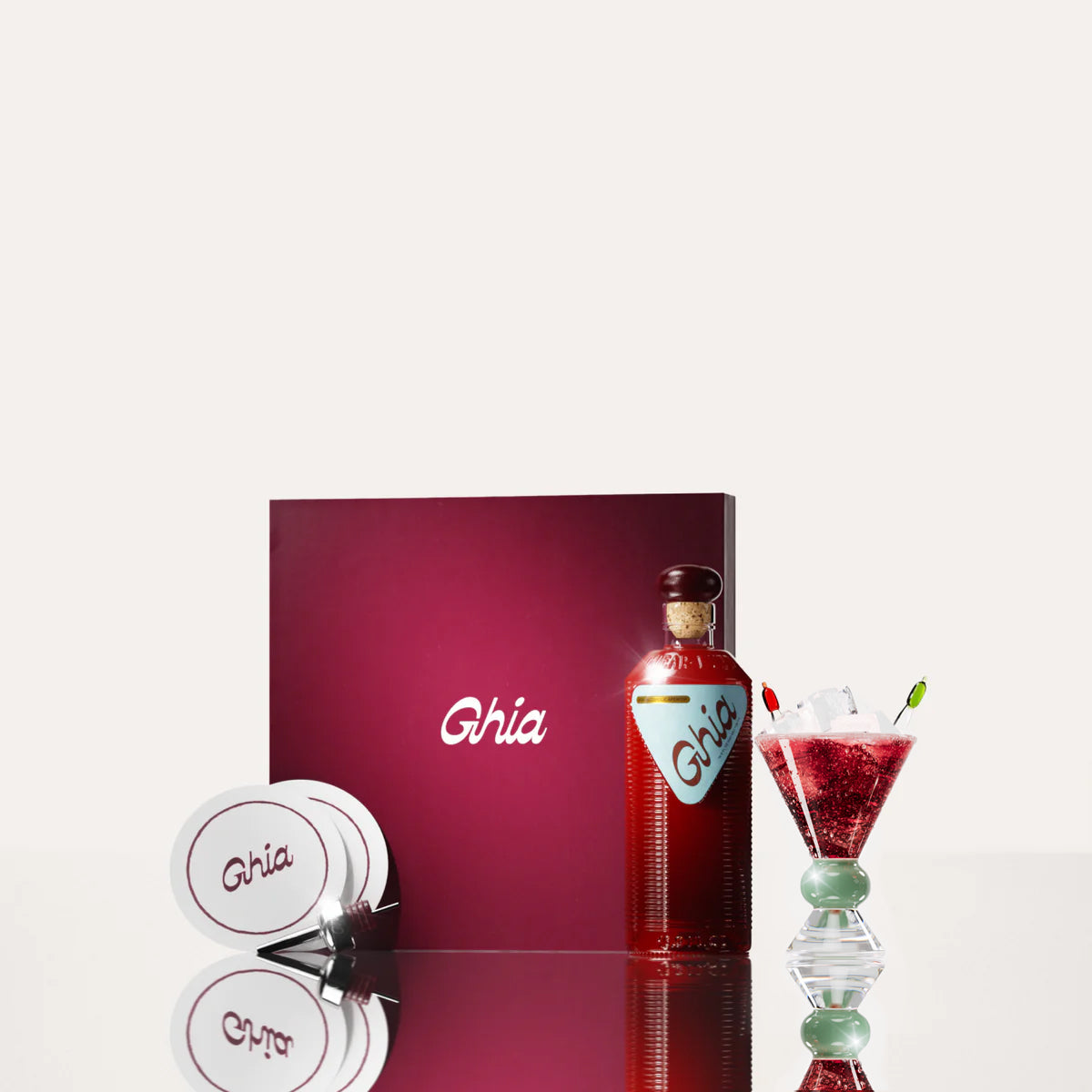 A Ghia non-alcoholic aperitif gift set with a burgundy box, paper coasters, 1 glass bottle of Original Apéritif, and a decorative cocktail glass that has a jade and clear base.