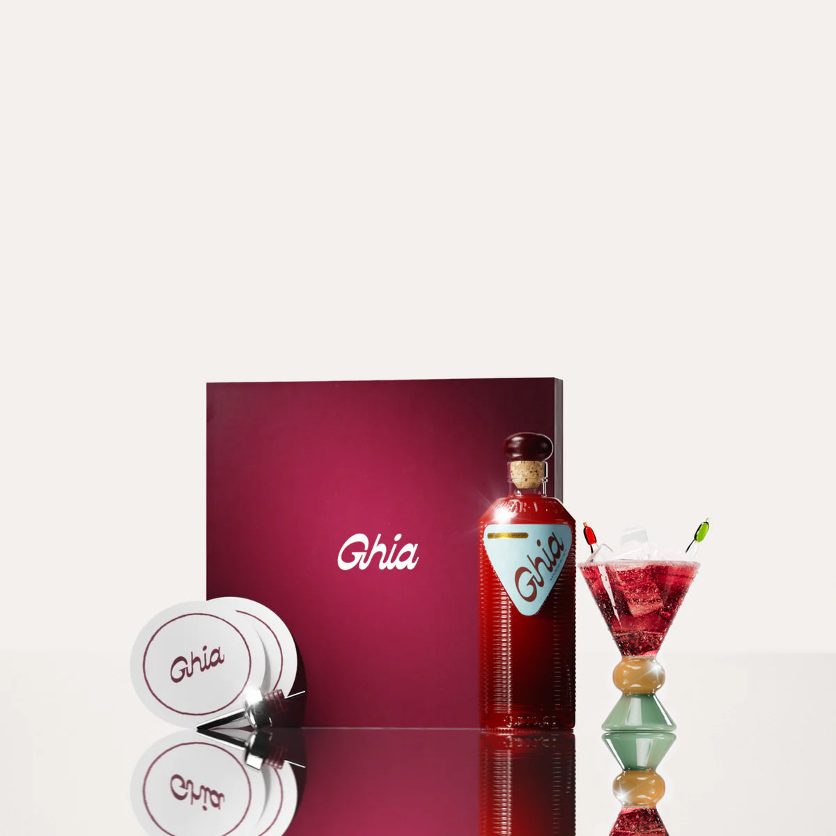 A Ghia non-alcoholic aperitif gift set with a burgundy box, paper coasters, 1 glass bottle of Original Apéritif, and a decorative cocktail glass that has a sand and jade base.