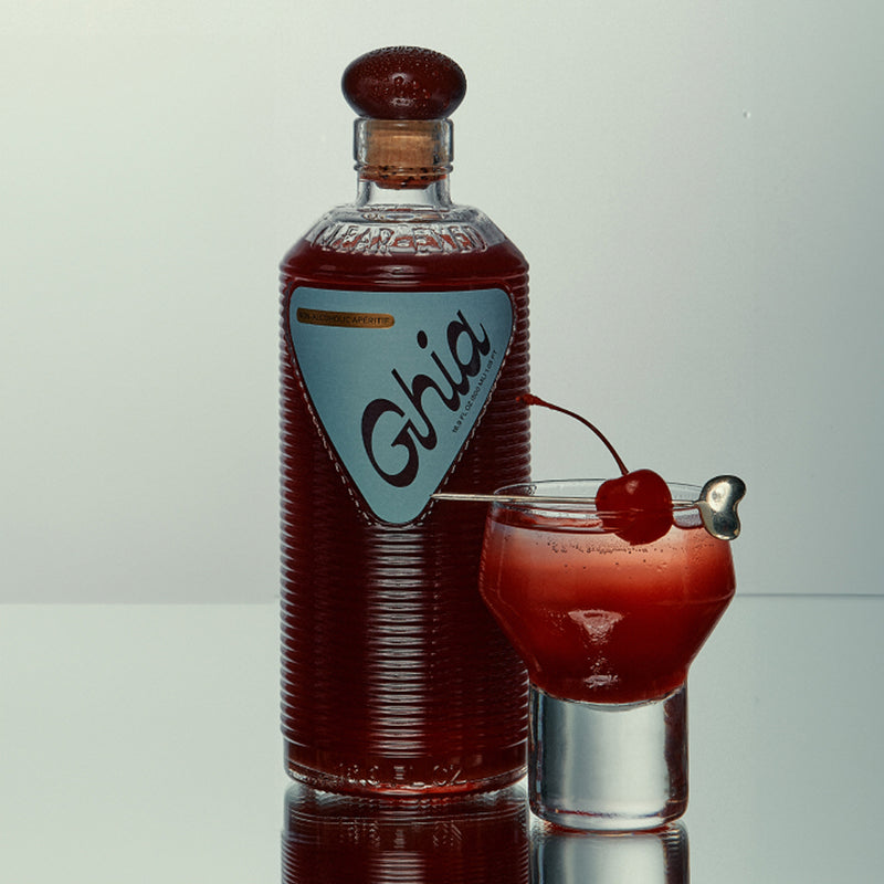 A bottle of Ghia non-alcoholic aperitif next to a glass of the drink, garnished with a cherry on a cocktail pick, reflecting a deep red color. The bottle has a blue label with "Ghia."