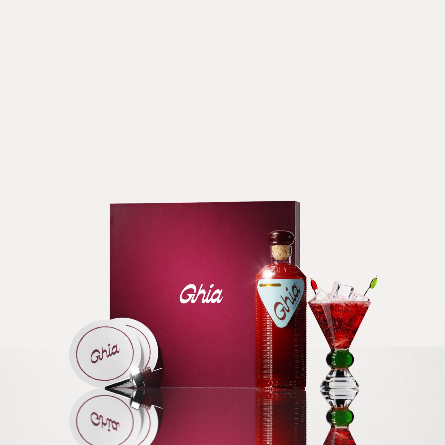 A Ghia non-alcoholic aperitif gift set with a burgundy box, paper coasters, 1 glass bottle of Original Apéritif, and a decorative cocktail glass that has a green and clear base.