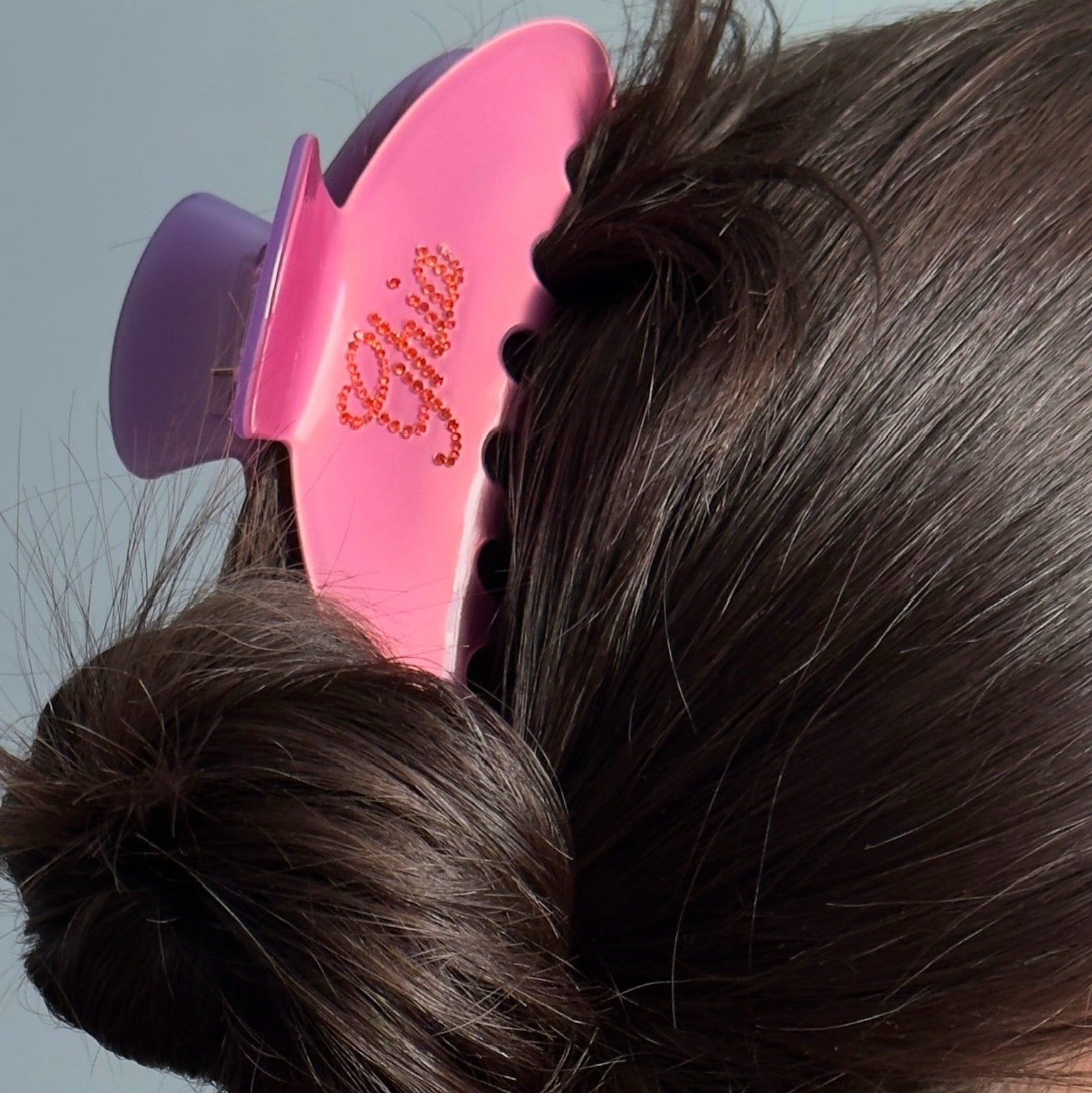 A close-up of dark brown hair secured with a pink and purple Ghia-branded claw clip, featuring decorative rhinestone lettering.