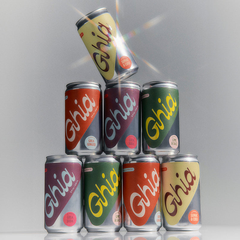 A stack of eight cans of Ghia non-alcoholic aperitif in various flavors: ginger, soda, lime & salt, sumac & chili, with bright, colorful labels and droplets of condensation on the cans.