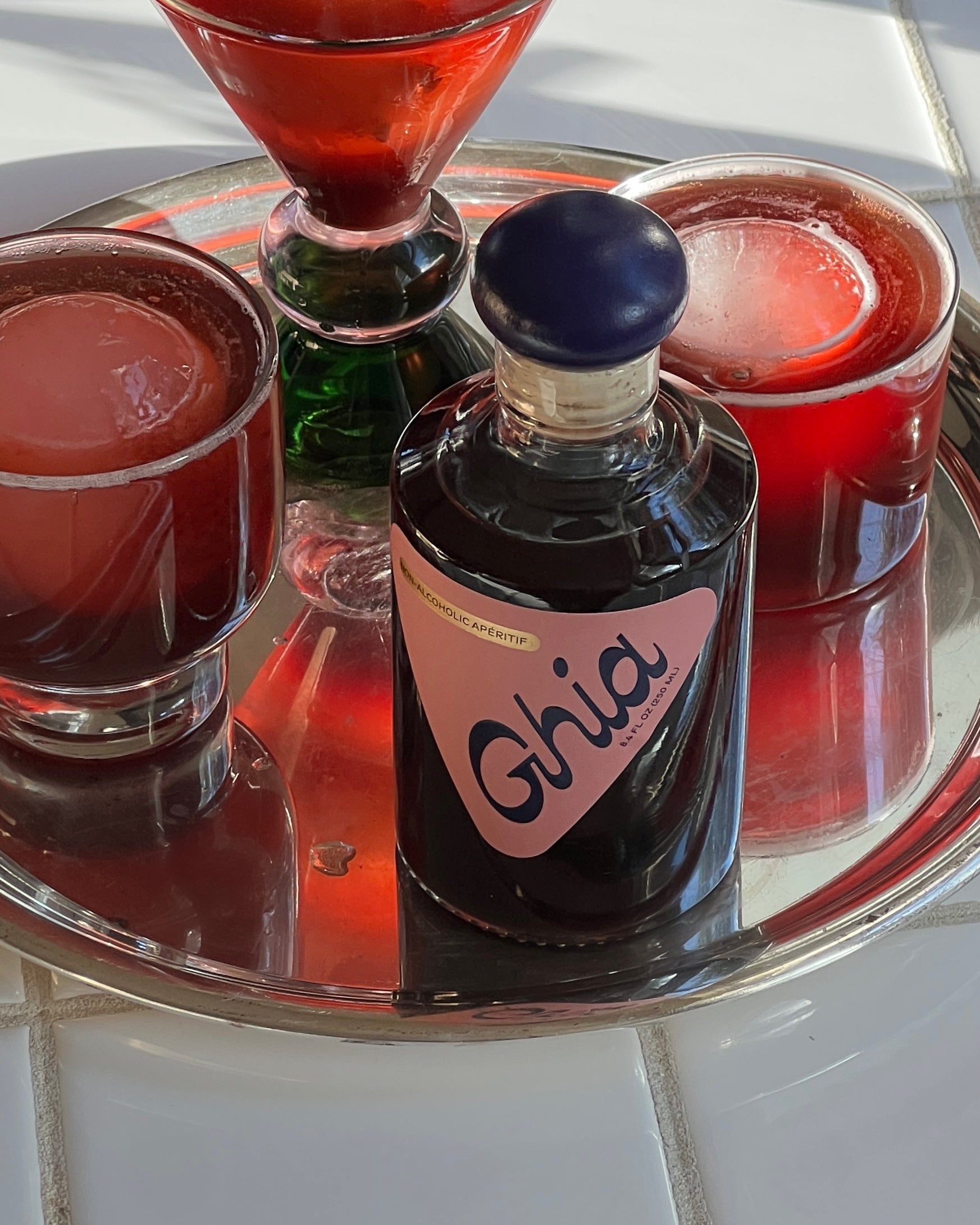 Dark glass bottle labeled 'Ghia' on a reflective tray with red drinks.