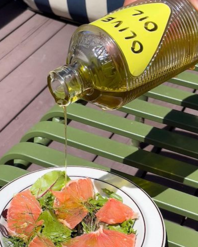 A ribbed glass bottle with a yellow label pours golden oil onto a fresh salad with grapefruit slices and greens, set on a green bench.