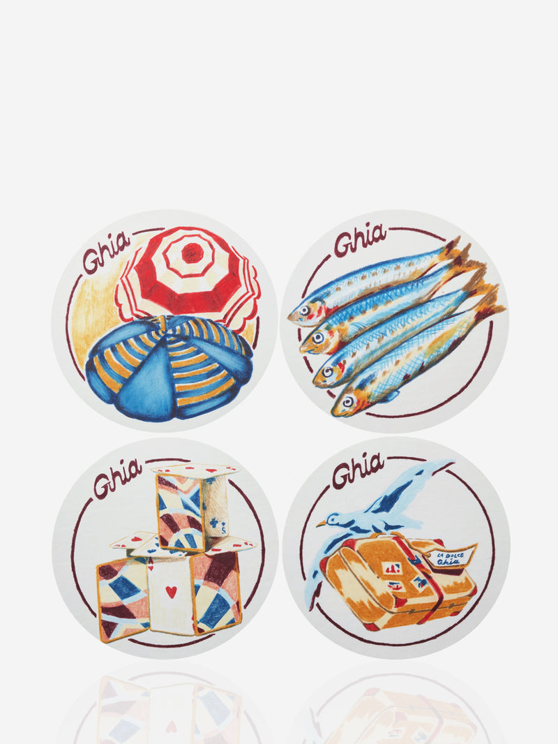 Four round Ghia coasters with illustrated designs featuring a beach umbrella, fish, stacked playing cards, and a suitcase with a bird.