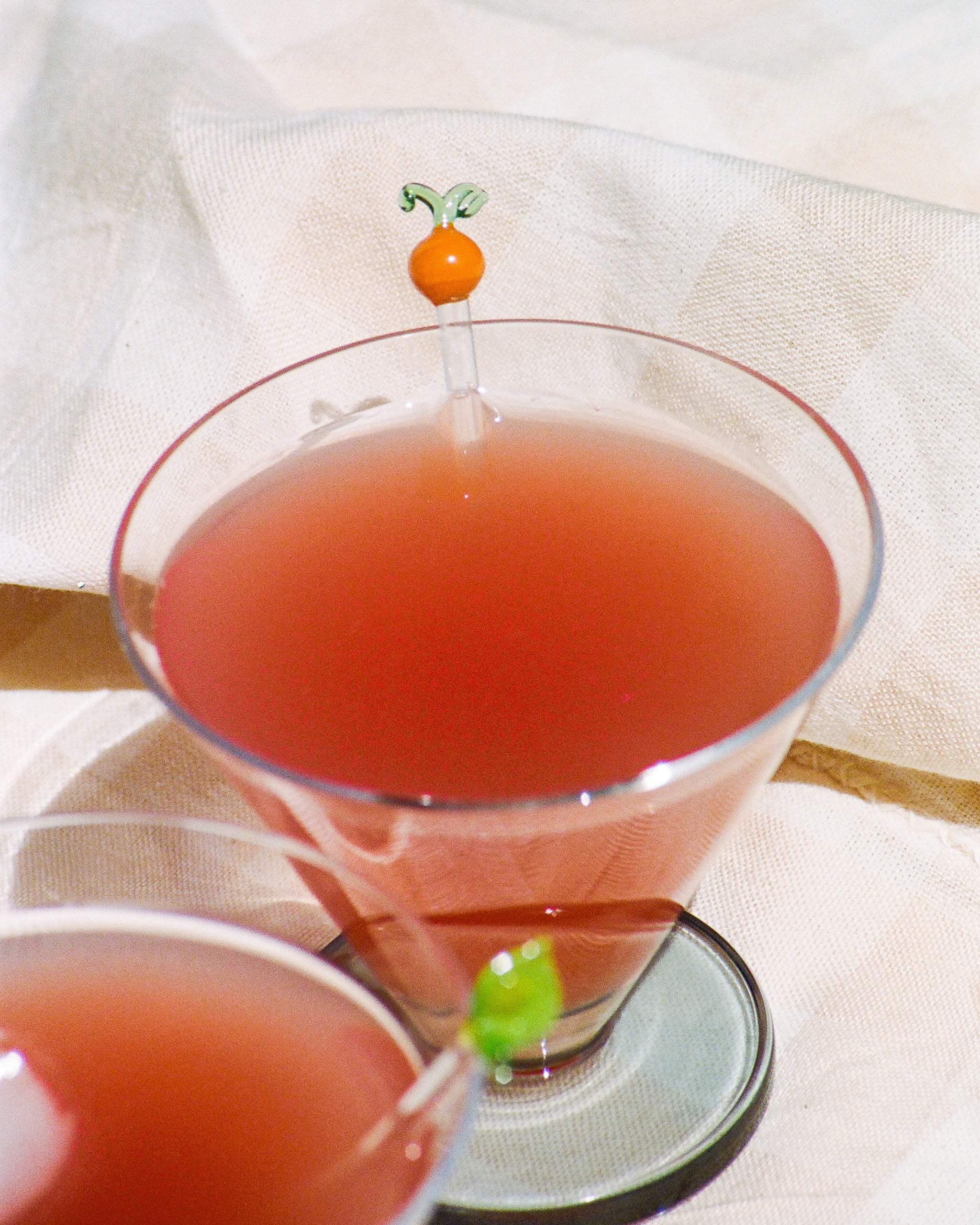 A close-up of two cocktail glasses filled with a pink drink, each garnished with a decorative stirrer featuring a tiny fruit-shaped top.