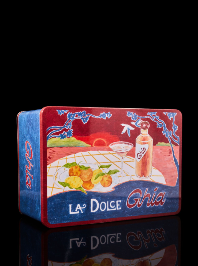 A colorful Ghia tin box featuring an illustrated scene with a bottle, cocktail glass, citrus fruits, and a sunset, labeled 